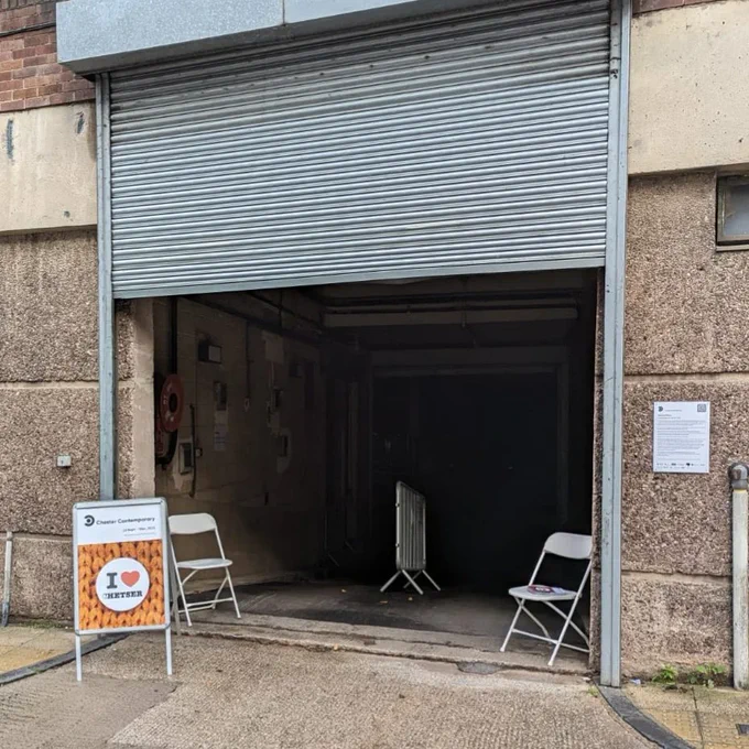 A picture of an industrial roller shutter entrance to a car park. Outside it is an a-board, tucked inside are two white folding chairs.
Image: © James Raine