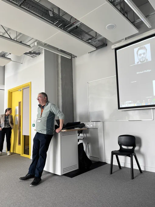 Man (John Gilligan) standing at top of room with projector with zoom on behind.