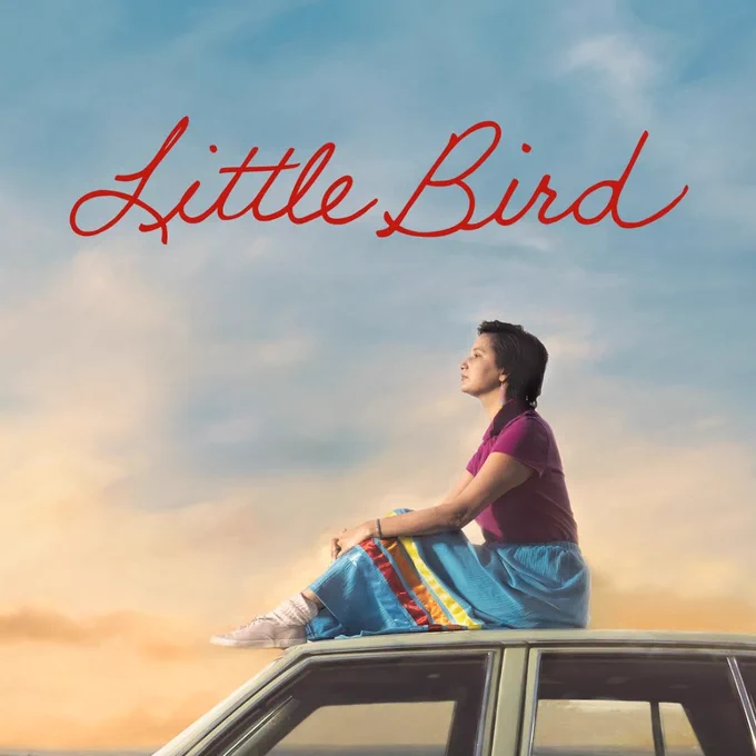 A young Indigenous woman with brown hair, wearing an traditional ribbon skirt and red shirt is sitting on top of a car in the sunset. Above her are the words "Little Bird."