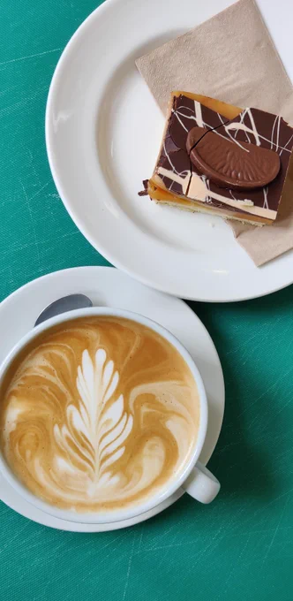A coffee in a white cup on a white saucer next to a plate containing a chocolate orange cake on a brown napkin.