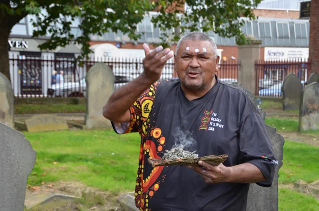 Mark stands in Warstone Lane Cemetery, he holds a piece of tree bark on which Rosemary is burning. His hand is raised to the smoke. 