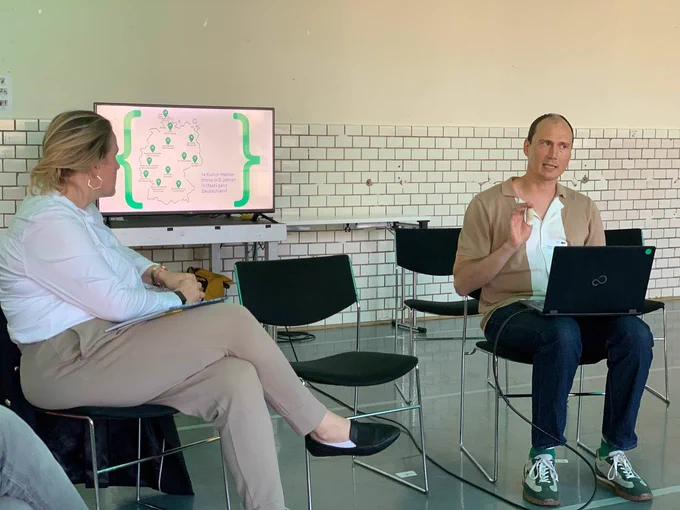 Foto vom Table Talk mit Philippe Genêt. Kurzhaariger Mann präsentiert mit Bildschirm und Laptop, am linken Bildrand sitzt eine Frau ihm zugewandt und hört zu