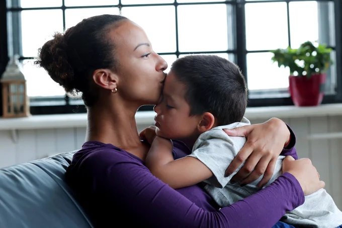 Adult and child sitting on a couch. The adult is hugging the child and kissing the child on the forehead.