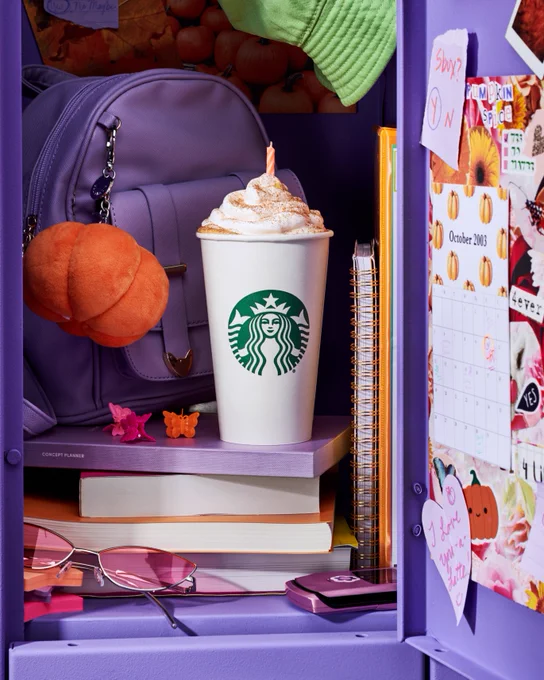 FRAME 1 ‘03 Locker: A Pumpkin Spice Latte with a lit birthday candle in the whip rests on stacked notebooks in an open purple school locker. Around it are other nostalgic items like a flip phone, pink sunglasses, butterfly clips, and a calendar that reads October 2003.