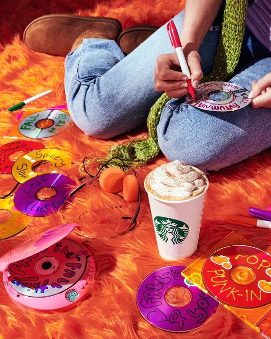 FRAME 2 ‘03 Mix Tapes: A Pumpkin Spice Latte with a lit birthday candle in the whip sits on a vibrant orange shag carpet. Behind it a person writes on a mix CD, with other CDs scattered around the scene that read “Pop-Punkin,” “Leaf (Get Out),” “Fall Mix ‘03,” and “Fall-ing 4 You.” 