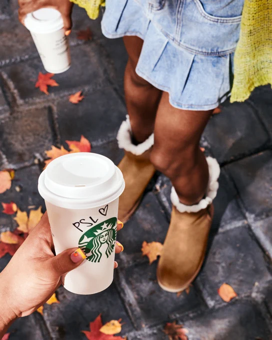 FRAME 3 ‘03 Fits: A person holds a Pumpkin Spice Latte with “PSL” and a heart sharpied on it against a street covered in fall leaves. Behind it we see a friend holding a PSL of their own wearing a denim skirt and fur lined boots.