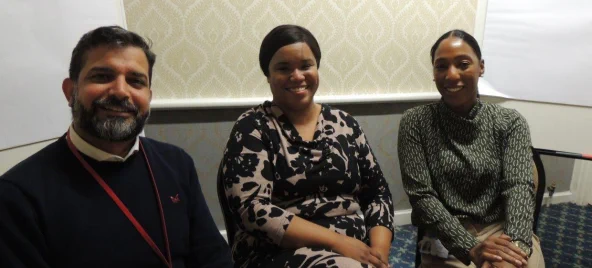 (left to right) Shakoor, Genine and Neddra are sitting down and  smiling at the camera. Shakoor is wearing a dark blue jumper, Genine is wearing a beige and black floral dress and Neddra is wearing a green and cream patterned top and beige trousers.