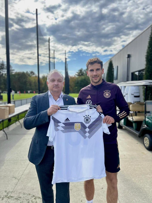 The deputy consul general stands next to Leon Goretzka as they both hold up a white jersey of the t mens national soccer team.