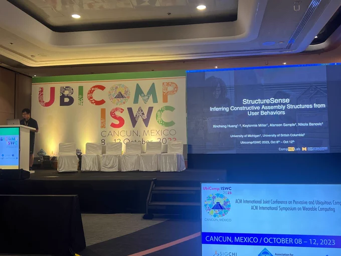 Xincheng presenting in front of a lectern. A sign reading UBICOMP / ISWC is behind the presenter, and the title slide is projected on a large screen in the right corner of the image.