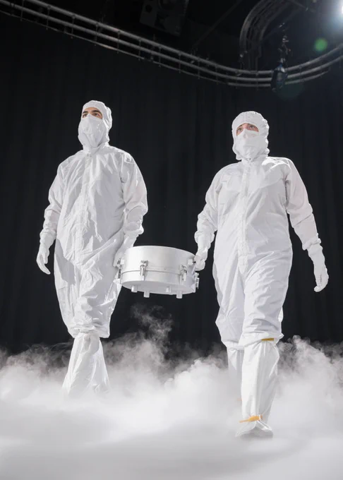 A staged photo shoot of the OSIRIS-REx container that was used to carry the Bennu asteroid sample. At NASA's Johnson Space Center, the participants are holding the container and are pictured in white clean room outfits. 