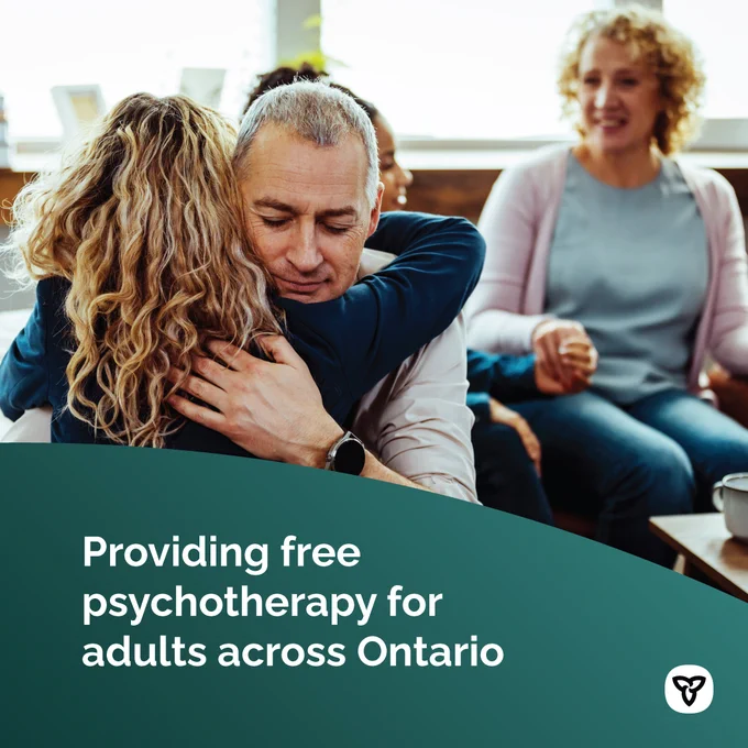 A man and woman hug during a group session, while two seated women converse in the background. Image text: Providing free psychotherapy for adults across Ontario.