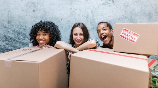 Three people looking excited standing behind and leaning against three large, cardboard boxes for moving house. A fragile sticker is stuck to one of the boxes.