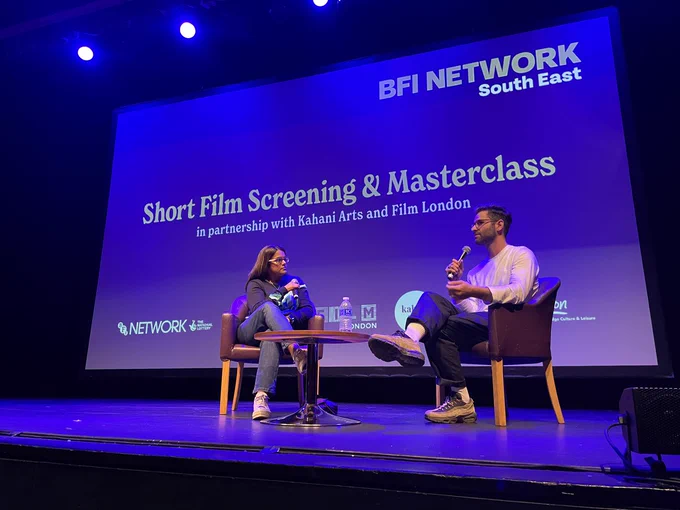 two people sitting on chairs on a stage talking into a microphone. Behind them is a projection saying short film screening and masterclass.