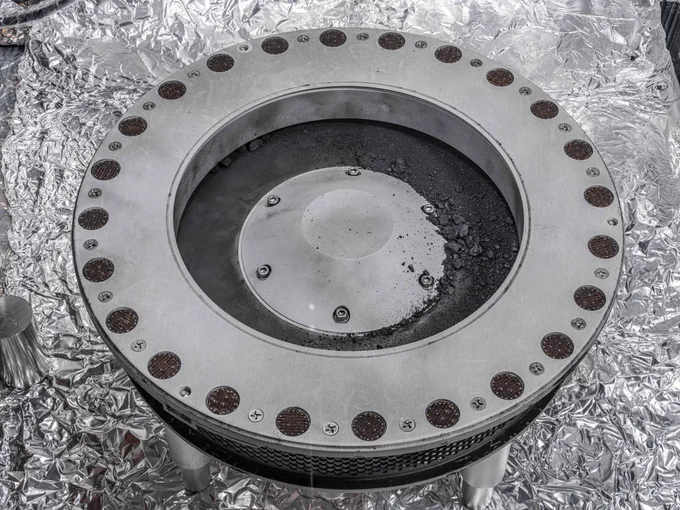 Circular metallic instrument of concentric circles on aluminum foil, the TAGSAM head from the OSIRIS-REx spacecraft. The outer ring contains many small circles containing small particles of black dust. The inner circle contains small rocks. Credit: NASA/Erika Blumenfeld & Joseph Aebersold