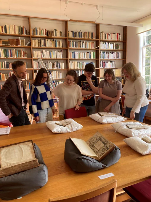 Six people stand in front of a desk which holds six white and grey book cushions. On each cushion is an old book, some held down using book snakes. Behind them is a wall of floor-to ceiling bookshelves.