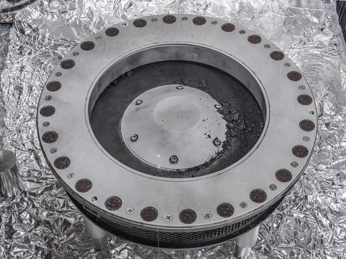 Circular metallic instrument of concentric circles on aluminum foil, the TAGSAM head from the OSIRIS-REx spacecraft. The outer ring contains many small circles containing small particles of black dust. The inner circle contains small rocks.  