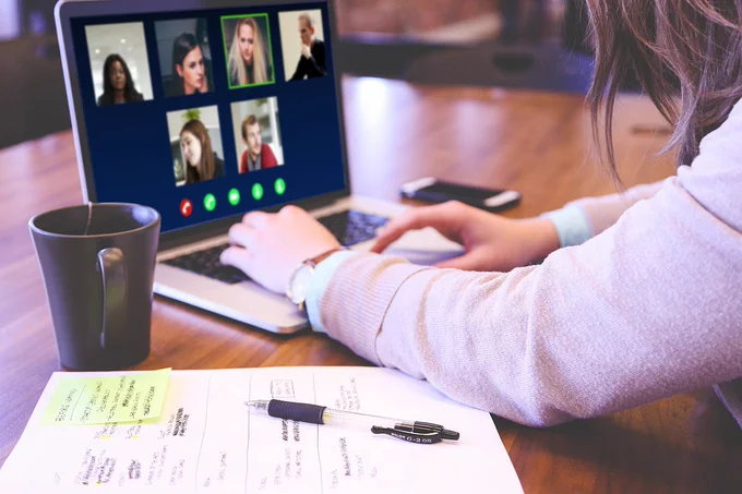 Person sat at a desk with a laptop open on a video call with multiple people. Cup with hot drink, pen and paper in the foreground.