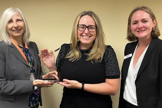 Photo of three people, one presenting an award to another, and all three smiling at the camera