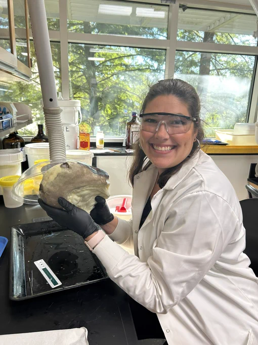 Lauren smiling in a lab coat and goggles with a sevengill shark head