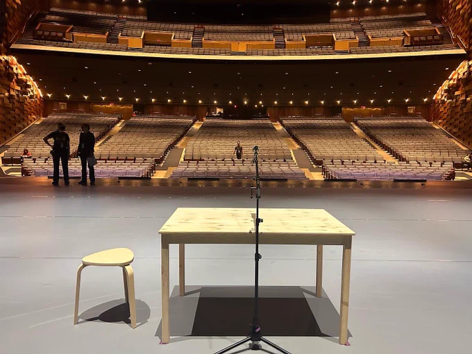 Photo of the Meridian Hall theatre in Toronto. The picture is take from the back of the stage and at the front, there is a microphone stand located in front of a rectangular, wooden table with a wooden stool on the left hand side. In the background are the theatre seats. Two people are on the left hand side of the stage and are looking away from the camera.