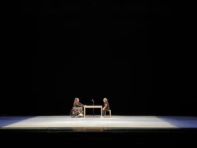 Dancers Maiya Leeke and Caroline Lofthouse on stage sitting at a wooden table and looking at each other. They are performing on a large grey stage against a black background.