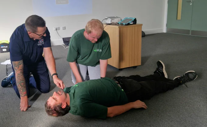A man in green lying on the floor. Two men looking at him.
