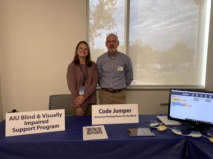 A woman and a man stand in front of a window and behind a table with many items on it, including desktop monitor, Code Jumper Pods, a piece of paper with a QR code on it, a sign reading "Code Jumper: American Printing House for the Blind," and another sign reading "AIU Blind & Visually Impaired Support Program."