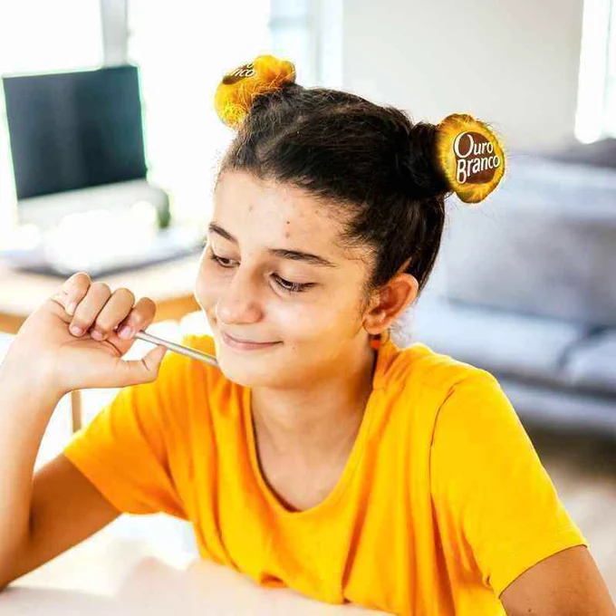 Imagem de uma menina com camiseta amarela e cabelo preso formando dois coques laterais, e em cada um deles temos uma embalagem de Ouro Branco, uma referência a brincadeira dos cabelos malucos das crianças nas escolas.