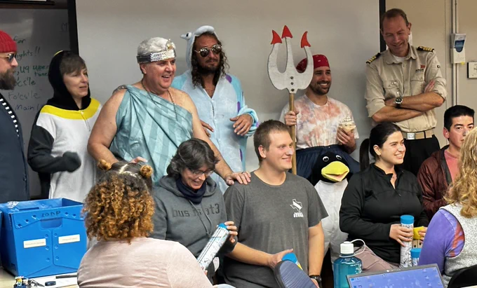 A group posing for a photo. The front row is kneeling. The back row has a person dressed as Davy Jones, a puffin, King Neptune, a spotted monster, a penguin holding Neptune's fork and the ship captain. 