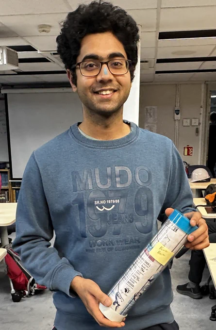 A person with glasses, dark skin and hair, standing and smiling and holding a document in a clear tube. His nose has a blue chalk streak.