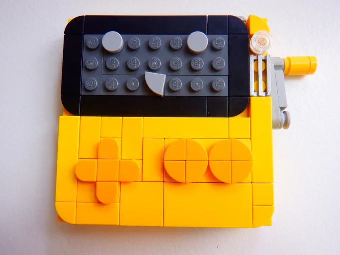 A photograph of a small and cute Playdate made from LEGO pieces. The body is yellow, the controls are there, the screen has a smiley face on it, the speaker is a little grill piece, it's very nice.