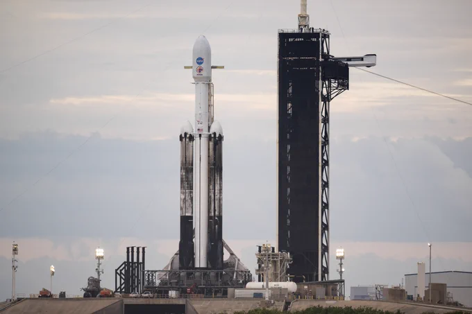 A SpaceX Falcon Heavy rocket with the Psyche spacecraft onboard is seen at Launch Complex 39A at NASA’s Kennedy Space Center in Florida. The tall, white rocket emblazoned with the blue NASA insignia and orange-and-purple Psyche mission branding is flanked by booster rockets smudged with black char from previous launches. A cloudy sky unfolds behind the launchpad. Credit: NASA/Aubrey Gemignani