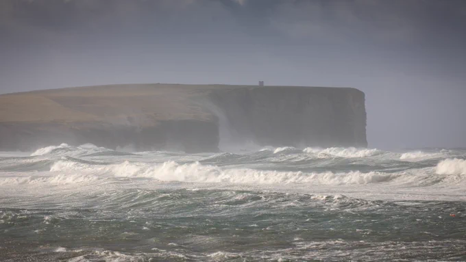 View towards Marwick Head with stormy seas in the foreground and sea spray shrouding part of the headland.
