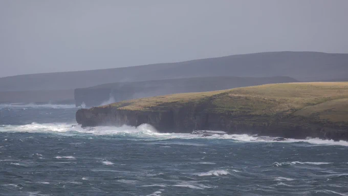 View along the Orkney coastline with headlands covered in sea spray and a dark grey sky above.