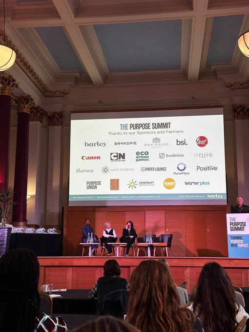 Photo of the stage at Purpose Summit. Screen shows the logos of partners and sponsors including Purpose Union.