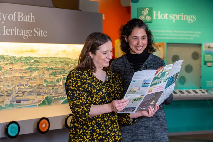 Two women are in the Bath World Heritage Centre looking at a map and smiling. 