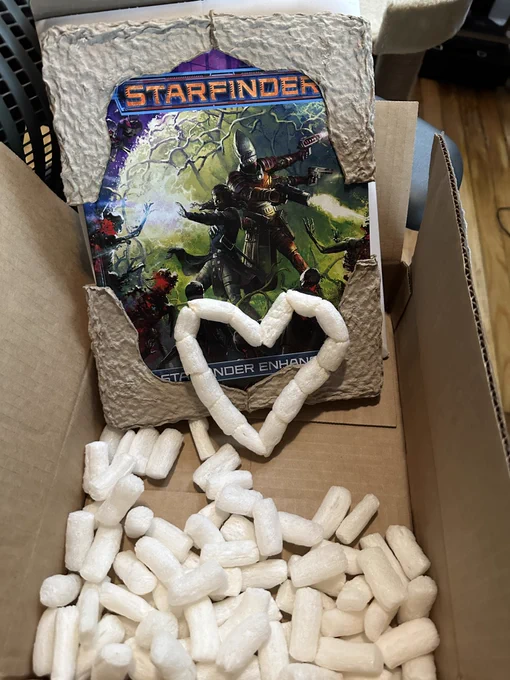 A hardcover copy of Starfinder Enhanced by Paizo, standing upright a cardboard shipping box, with cardboard corner protectors in place on the book's corners. The bottom of the box is littered with starch packing peanuts, and a heart shape made from peanuts stuck together is leaning against the cover of the book.