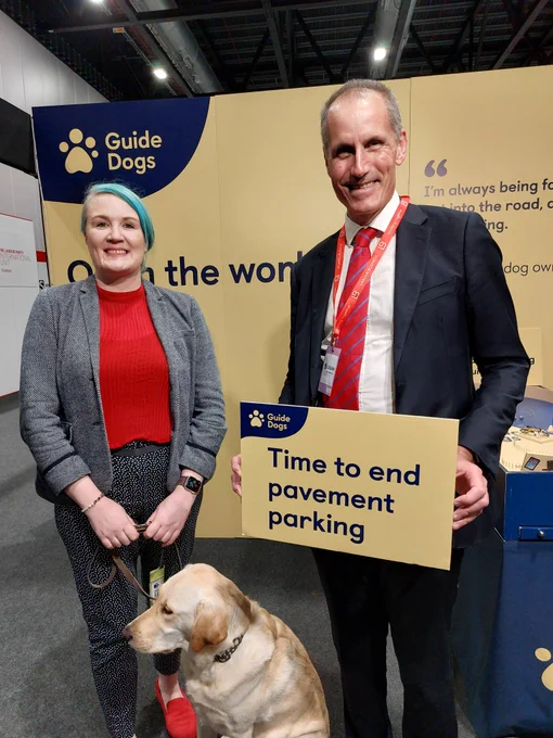 A man and a woman in front of a yellow backdrop. The woman is a guide dog owner. The man is holding a yellow sign saying "Time to end pavement parking".