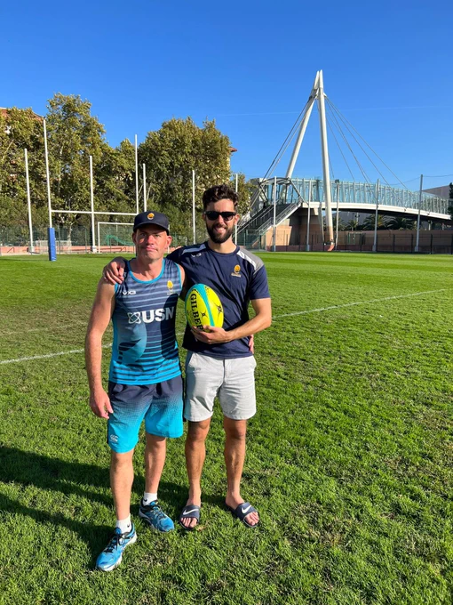 Photo shows Matt lancett and chris styles standing linking arms holding a VI rugby ball, on a grass rugby pitch with rugby posts in background and blue sky