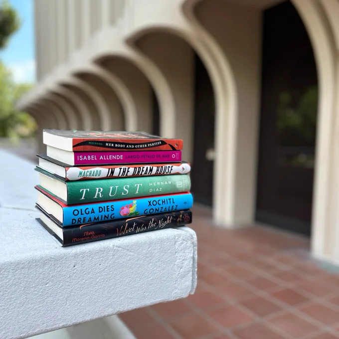 A stack of books by Latino/Hispanic authors with Langson Library blurred in the background.