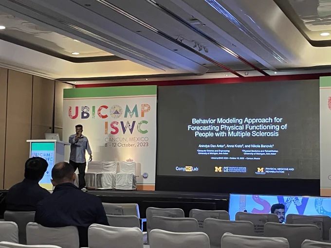 Anindya presenting in front of a lectern. A sign reading UBICOMP / ISWC is behind the presenter, and the title slide is projected on a large screen in the right corner of the image.