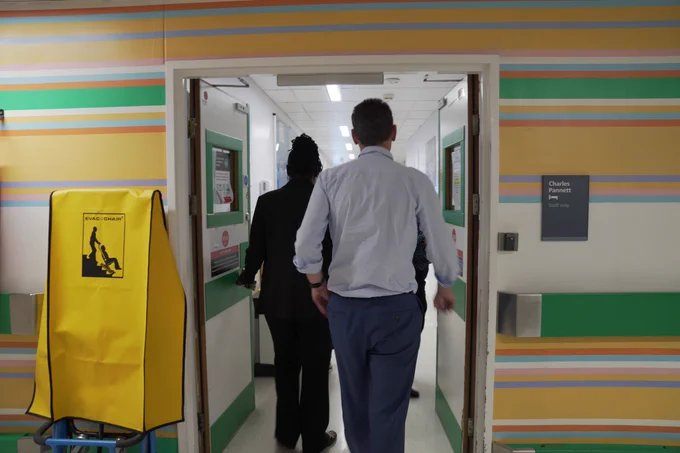 Image shows two NHS England staff walking through a hospital ward at Imperial College Healthcare.
