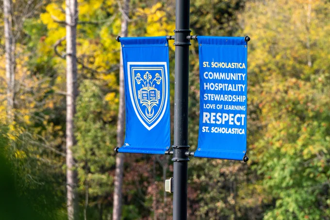 Two St. Scholastica banners on pole with fall trees in the background. The left banner features the St. Scholastica shield and the right banner features text "St. Scholastica, Community, Hospitality, Stewardship, Love of Learning, Respect, St. Scholastica".