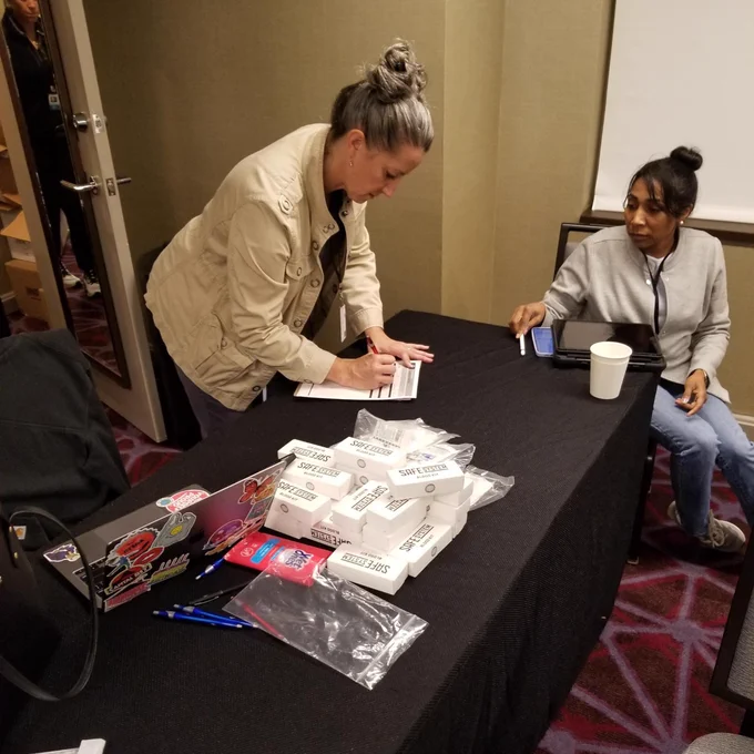 Woman standing and writing on paper while another woman sits beside her at table.