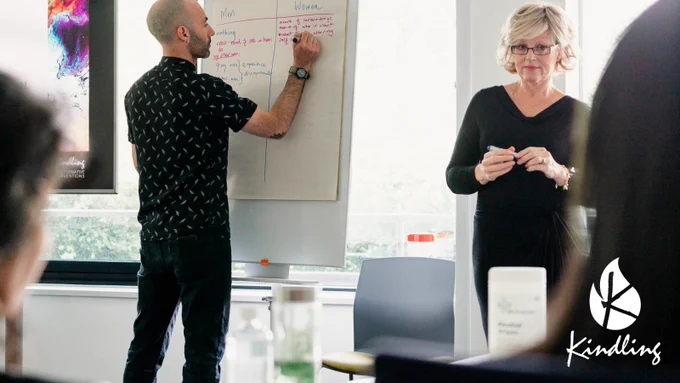 Nate and Rachel, two white adults wearing smart black clothes, are at the front of a room of people, leading a training session. At the bottom right corner is the word Kindling with a white flame icon above it, forming the full Kindling logo.