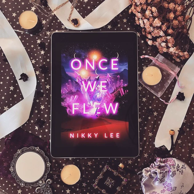 A book cover shown on an iPad. The cover has a landscape with purple crystals, several moons, and a sand-sailing craft. Its title is Once We Flew, and the author is Nikky Lee.

The book is laid on a starry background. Around it are tealights, ribbon, dried flowers, and ornate trinkets, all in pink, purple, white, and cream tones. The objects are scattered about to feel like a cluttered desk.