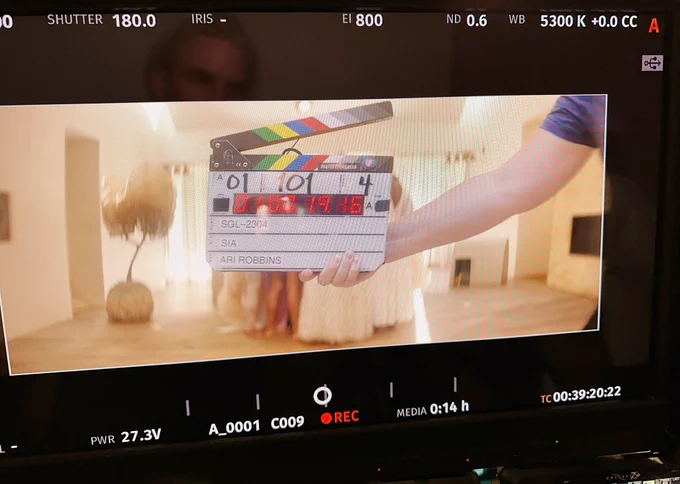 A BTS pic of the "Gimme Love" music video with a clapperboard in front of a group of people in all white
