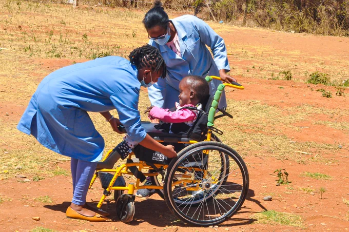 Two women in blue coats adjust a young girl in a yellow wheelchair.