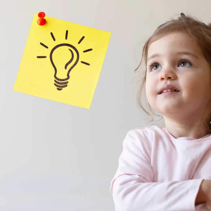 Stock photo of a toddler girl with light brown hair and light brown eyes, looking up towards a sticky note with a light bulb drawn on it. 