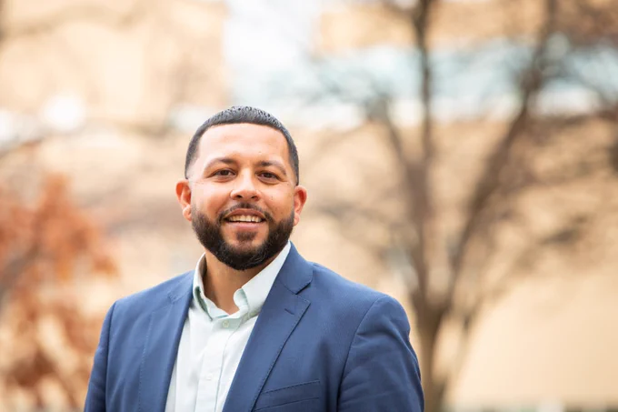 A professional photo of Prof. Velazquez. He is wearing a blue suit jacket and is outside, with the background artistically faded.